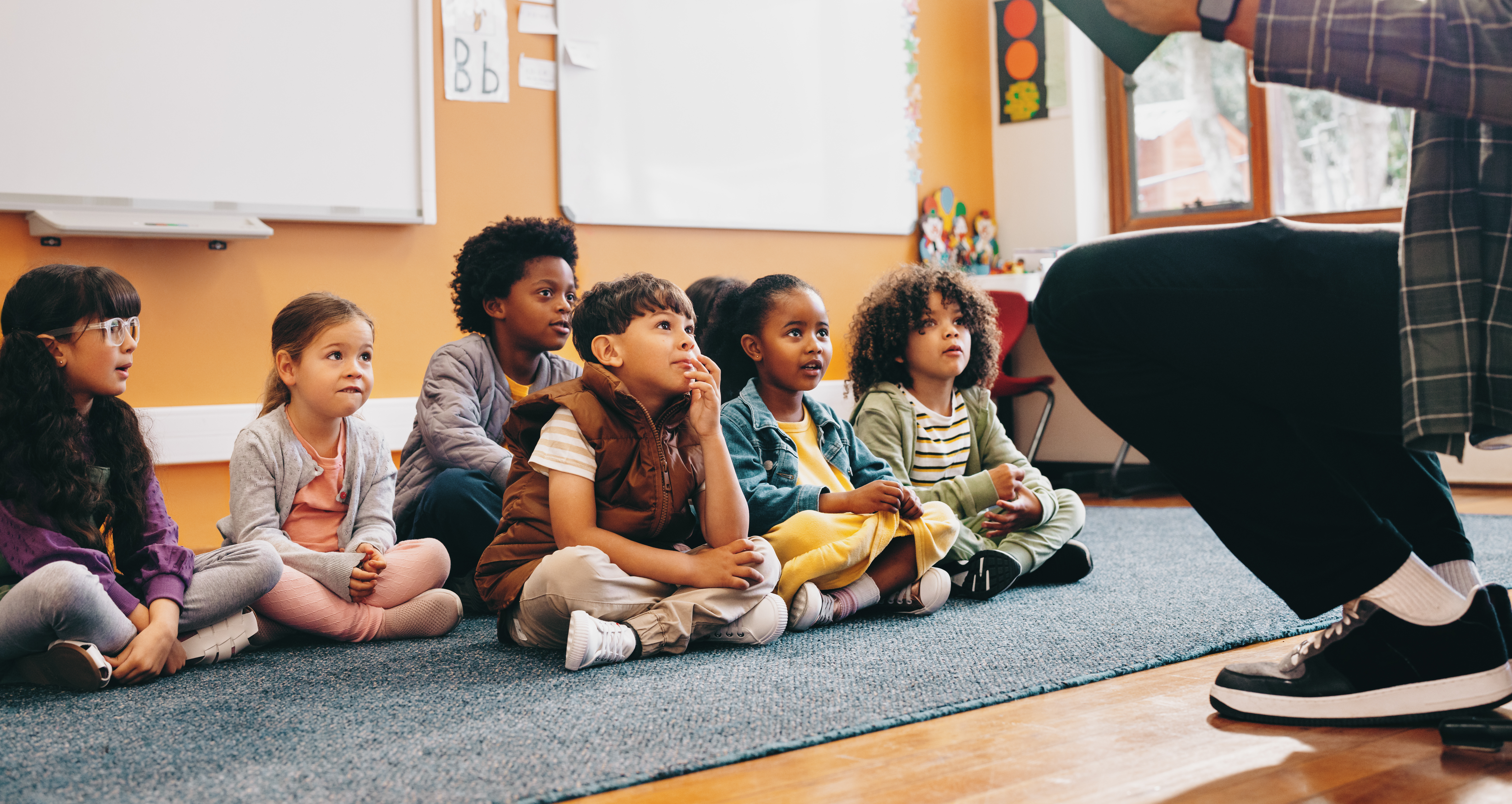 Elementary school students sit on a carpet and listen to a story from their teacher.